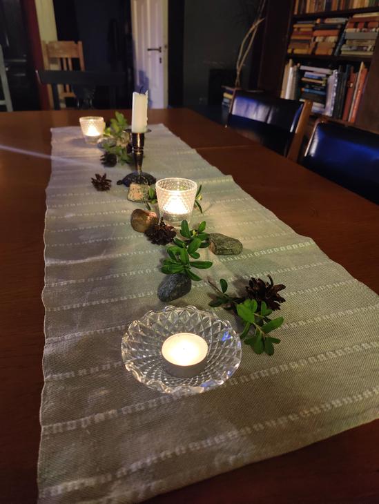 Kotikirjaston tumma puupöytä, jonka päällä pellavaliina, kynttilöitä, männynkäpyjä ja puolukanvarpuja