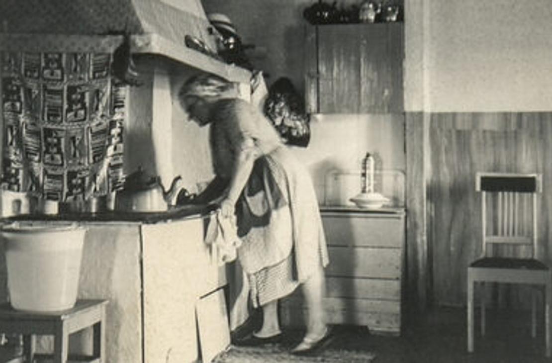 Mustavalkoinen valokuva, jossa nainen puuhellan äärellä.