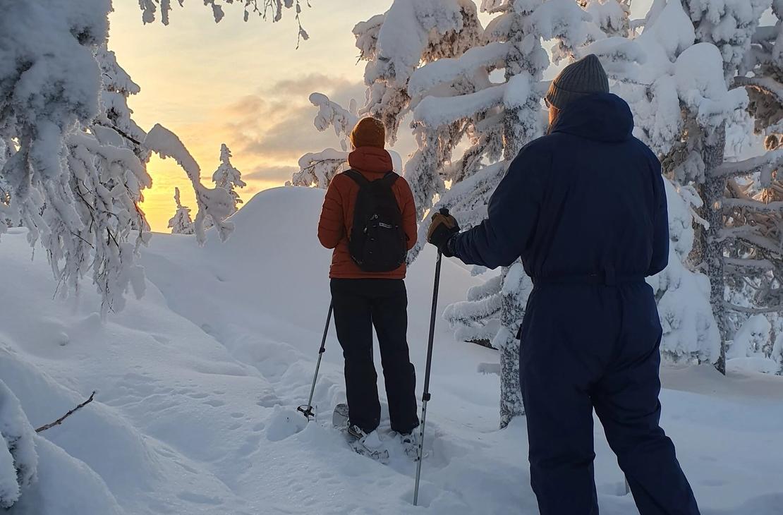 Kaksi lumikenkäilijää lumisessa metsässä, auringonlasku värjää horisontin keltaiseksi