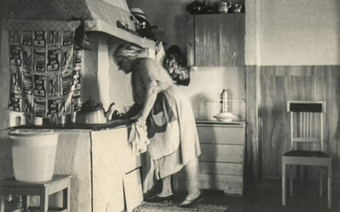 Mustavalkoinen valokuva, jossa nainen puuhellan äärellä.