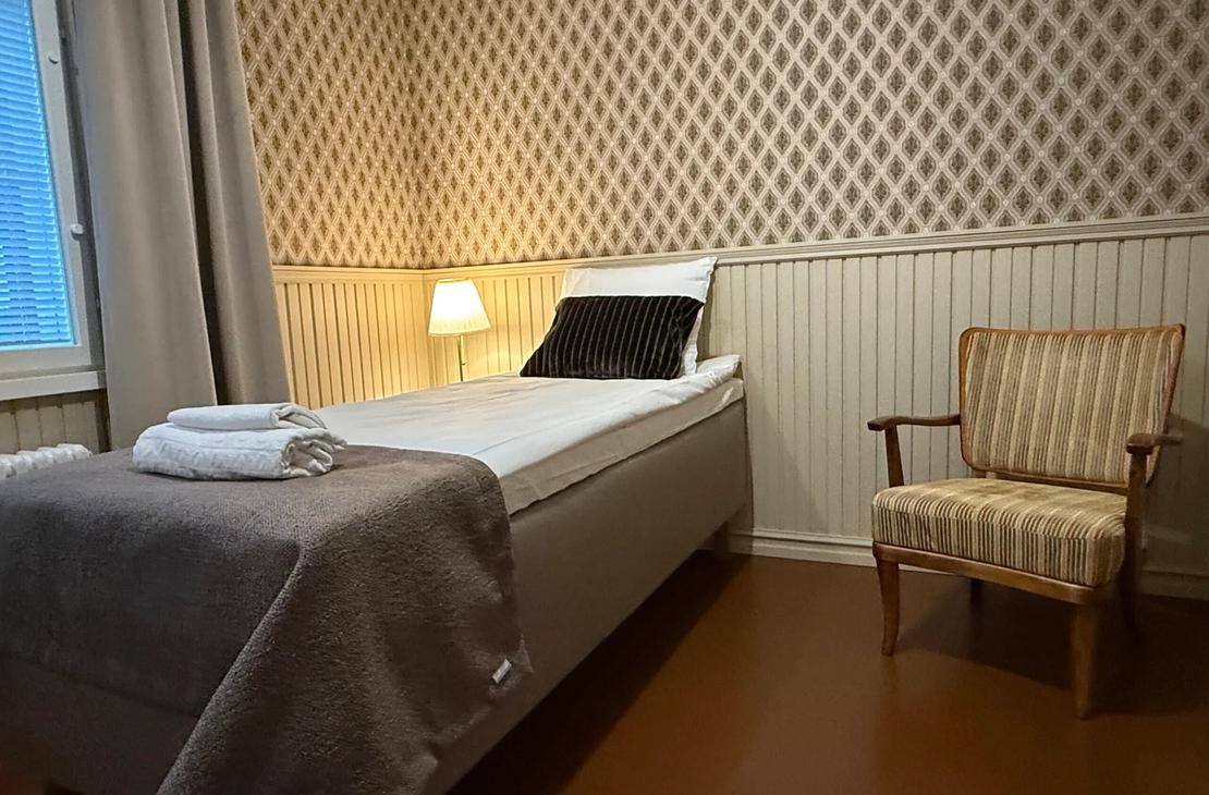 Sänky, lukuvalo ja nojatuoli huoneessa, jossa punertava tapettiseinä ja puiset puolipaneelit.