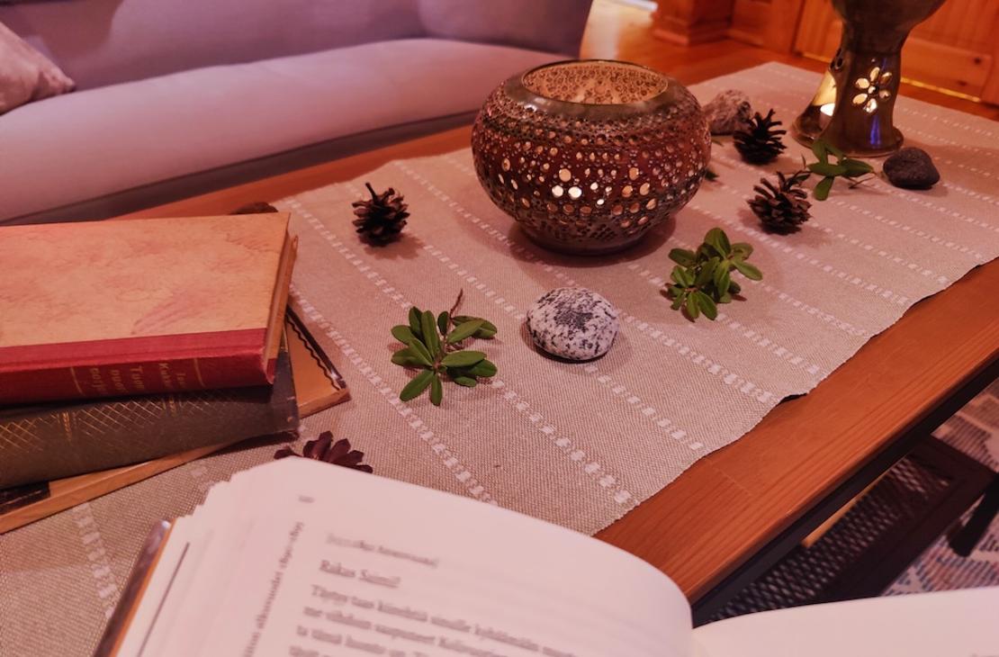 Kotikirjaston puupöytä, jonka päällä pellavaliina, kynttilöitä, männynkäpyjä ja puolukanvarpuja