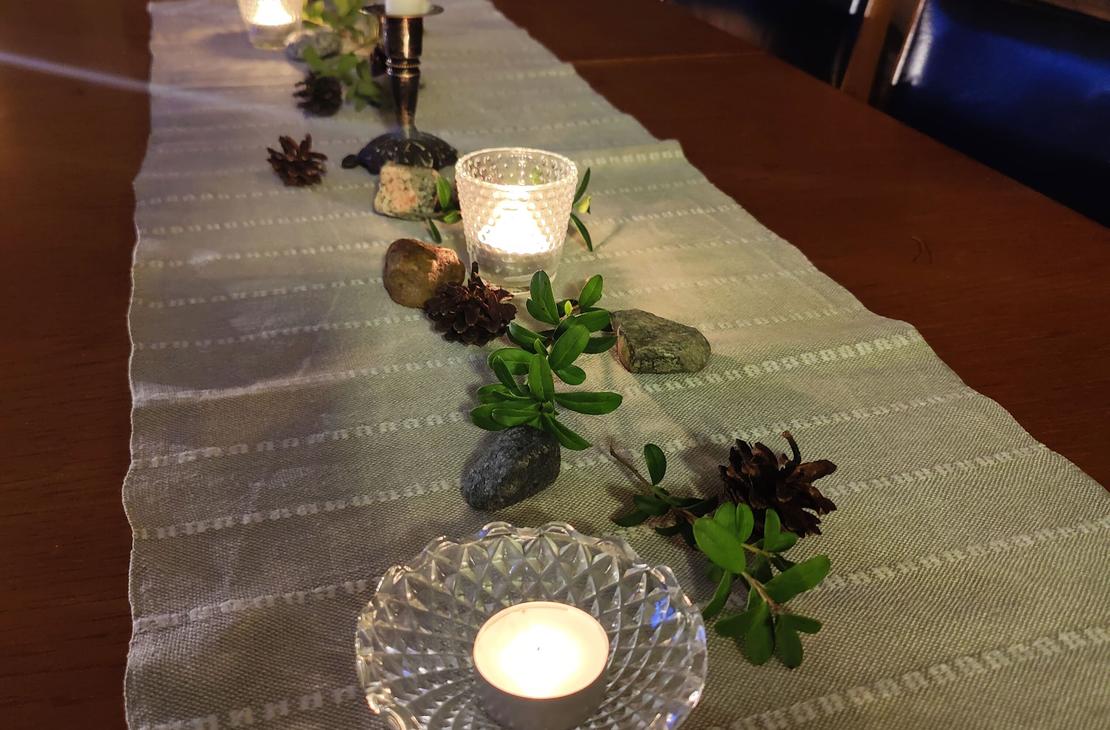Kotikirjaston tumma puupöytä, jonka päällä pellavaliina, kynttilöitä, männynkäpyjä ja puolukanvarpuja