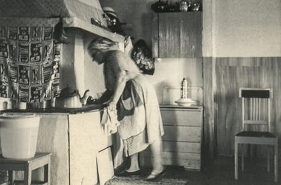 Mustavalkoinen valokuva, jossa nainen puuhellan äärellä.