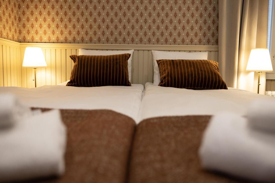 Kaksi sänkyä kuvattuna jalkopäästä; sängyn päädyssä punertava tapettiseinä ja puiset puolipaneelit.