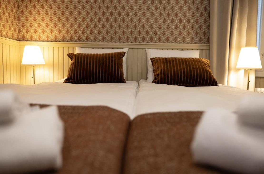 Kaksi sänkyä kuvattuna jalkopäästä; sängyn päädyssä punertava tapettiseinä ja puiset puolipaneelit.