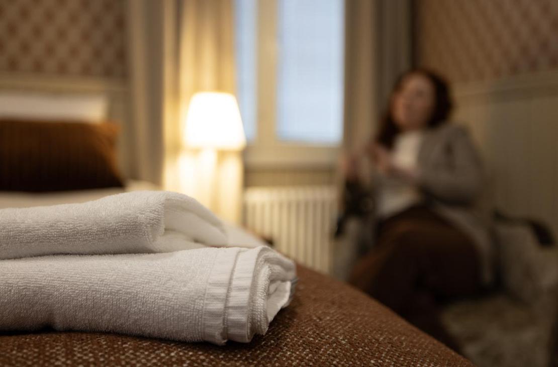 Kaksi pyyhettä aseteltuna sängylle etualalle; taustalla sumeana näkyvä henkilö istuu nojatuolissa huoneen rauhallisessa, vaalean ja punaruskean sävyisessä sisustuksessa.