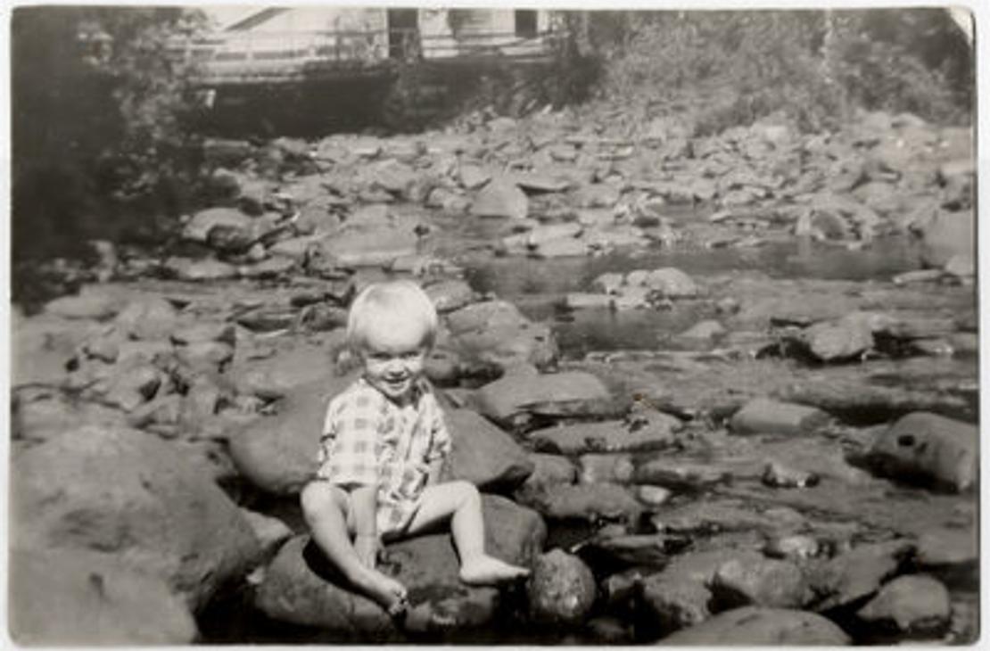 Mustavalkoinen kuva, jossa lapsi istuu kesäasussa myllyn rantakivillä
