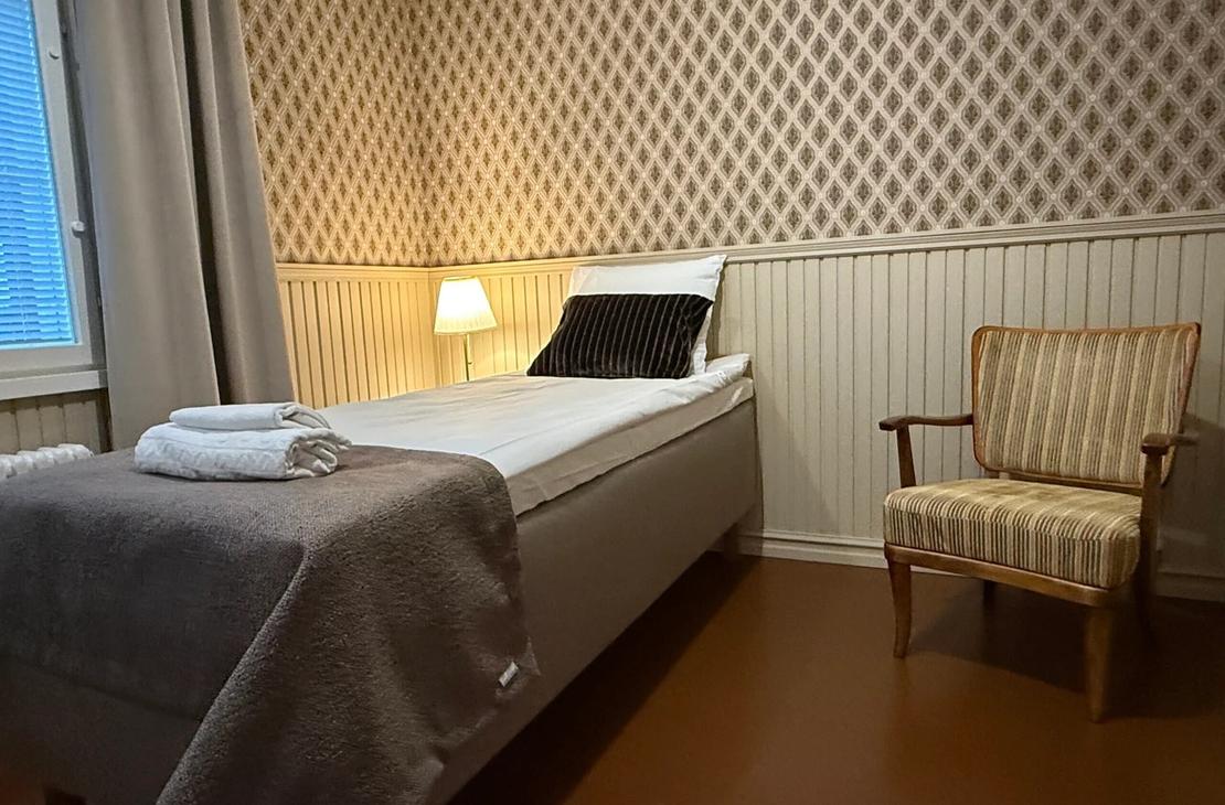 Sänky, lukuvalo ja nojatuoli huoneessa, jossa punertava tapettiseinä ja puiset puolipaneelit.