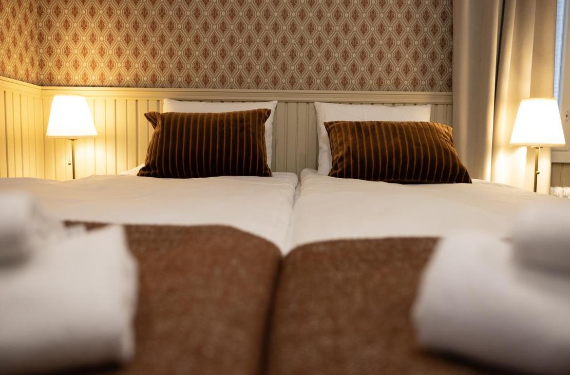 Kaksi sänkyä kuvattuna jalkopäästä; sängyn päädyssä punertava tapettiseinä ja puiset puolipaneelit.