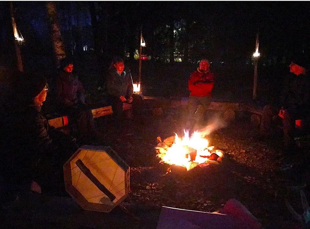 Ihmisä nuotion ympärillä illan pimeydessä nuotion ja kynttilöiden valaistessa vain hieman