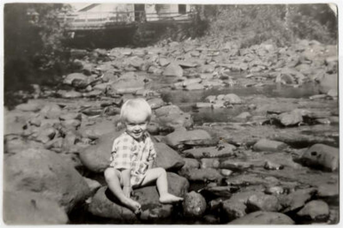 Mustavalkoinen kuva, jossa lapsi istuu kesäasussa myllyn rantakivillä