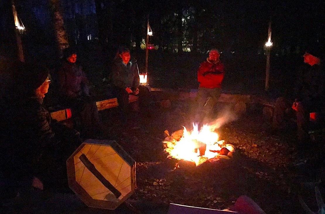 Ihmisä nuotion ympärillä illan pimeydessä nuotion ja kynttilöiden valaistessa vain hieman