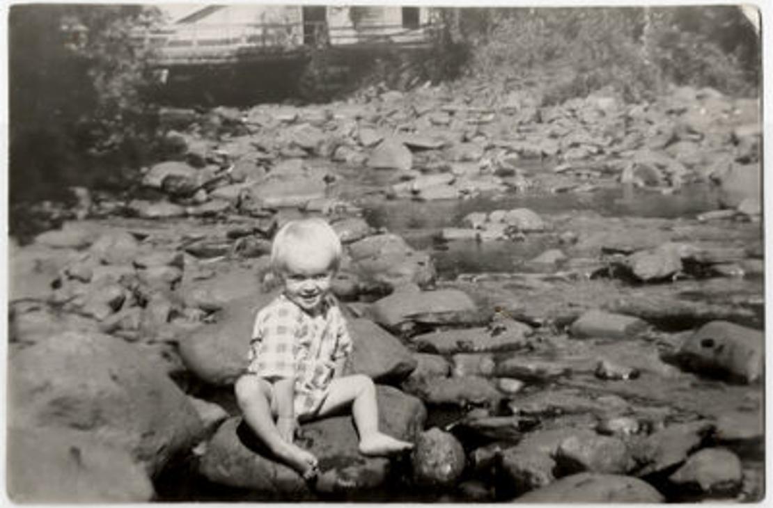 Mustavalkoinen kuva, jossa lapsi istuu kesäasussa myllyn rantakivillä